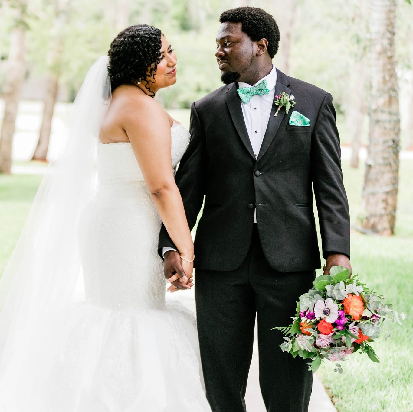 man wearing black tuxedo and seafoam accessories holding the bridal bouquet while standing next to his bride in a mermaid gown and veil