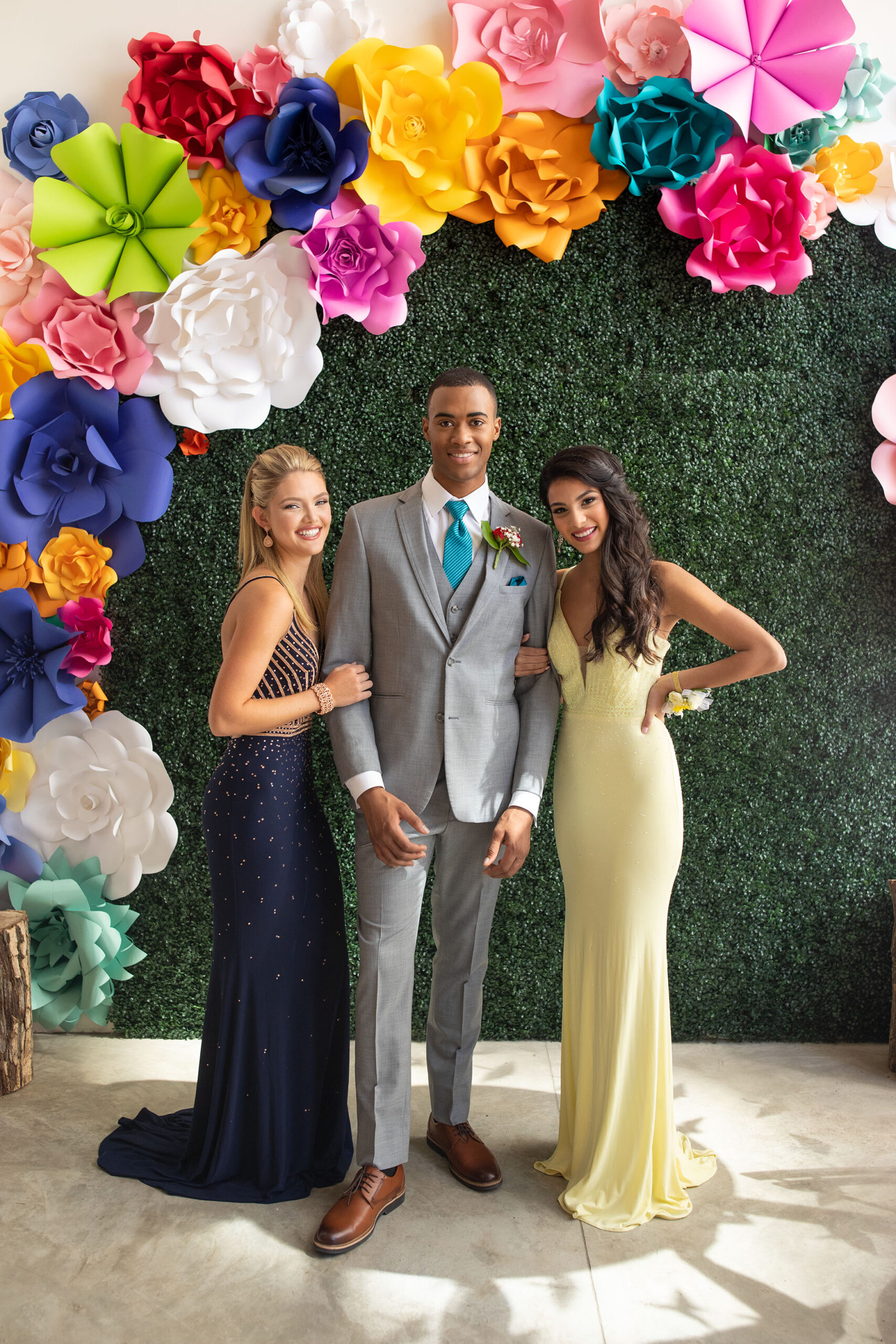 prom after-party picture with guy in grey suit, blonde female in navy dress and brunette in yellow dress standing in front of moss covered wall with brightly colored paper flowers