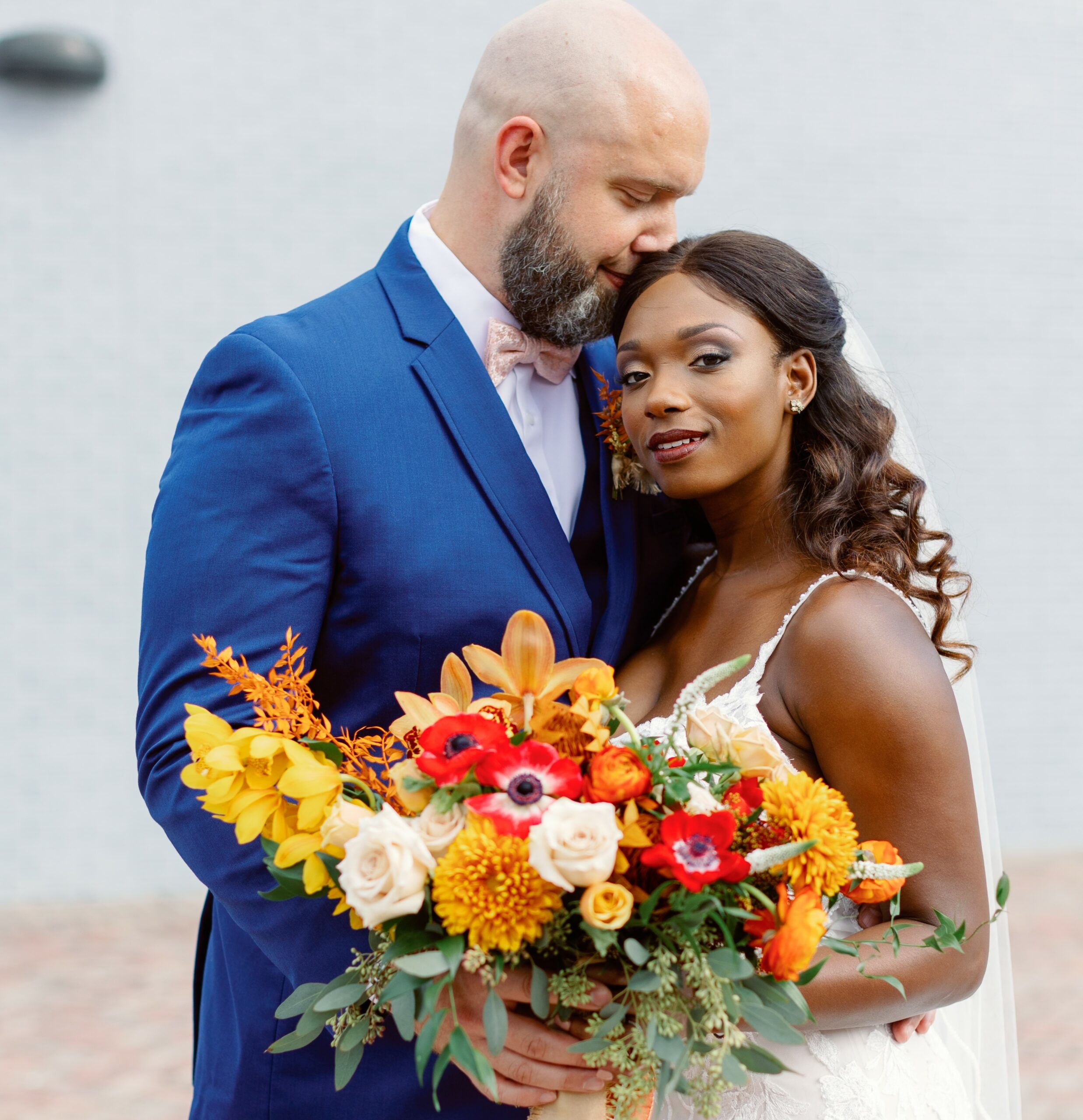 black bride in white dress holding colorful bouquet while standing next to white groom in blue suit