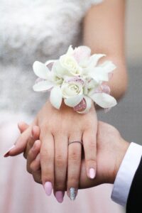 save money on prom - man holding hand of lady with pink and silver painted nails and a wrist corsage with white flowers and pink ribbon