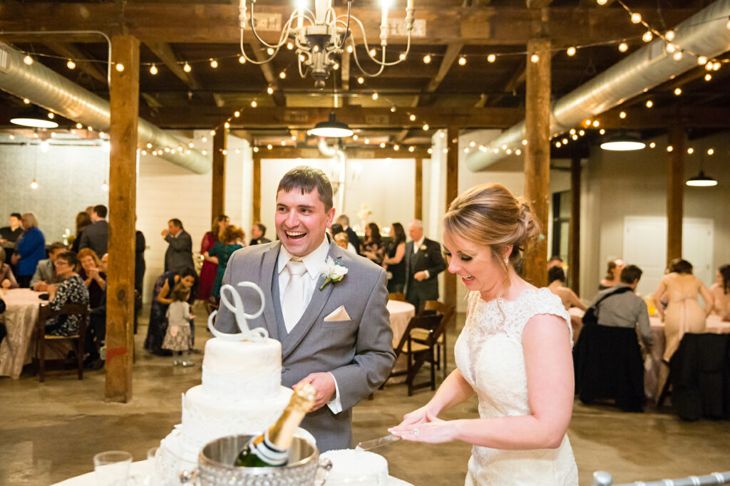 anniversary reception - groom in grey tuxedo standing next to bride wearing lace dress cutting the three tier wedding cake with their guests watching in the background