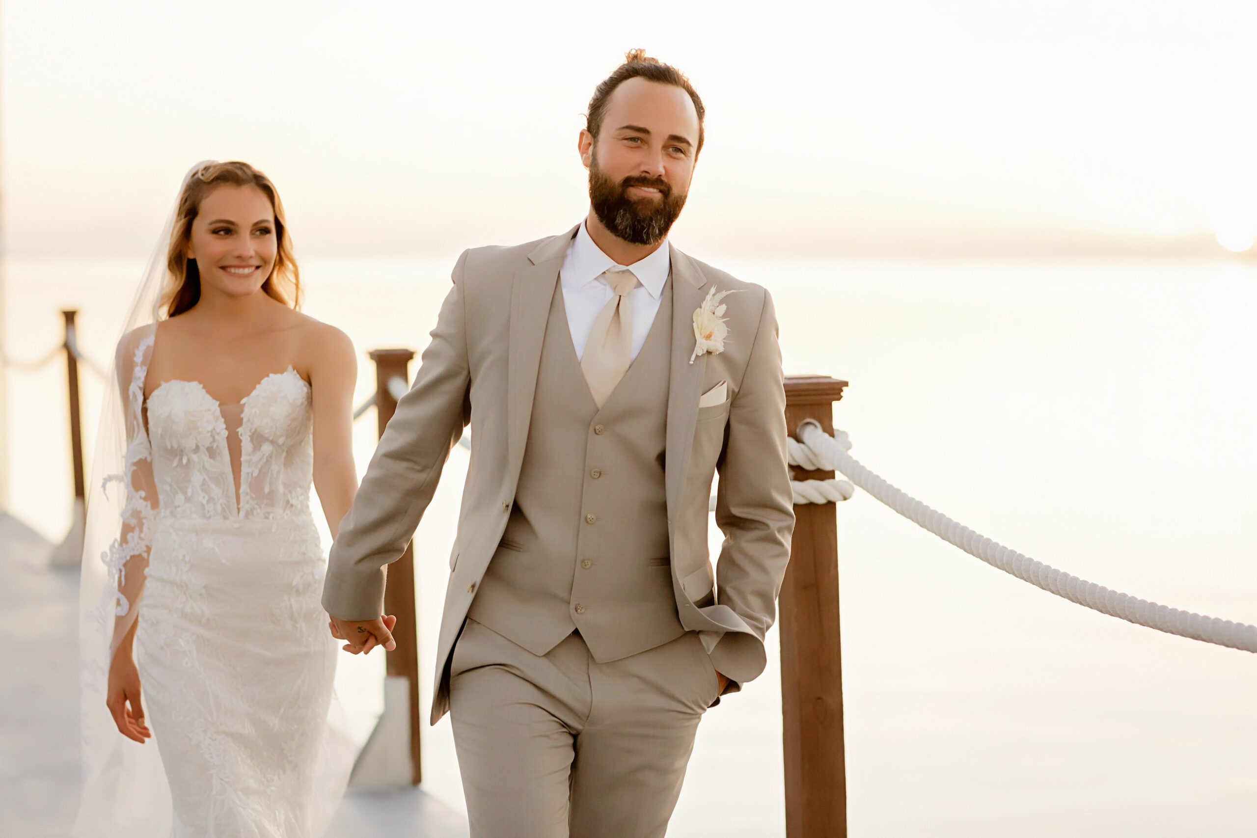 bride and groom wearing tan suit walking along a peer on the waterfront hand in hand