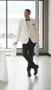 man in ivory shawl dinner jacket and black pants standing next to chair