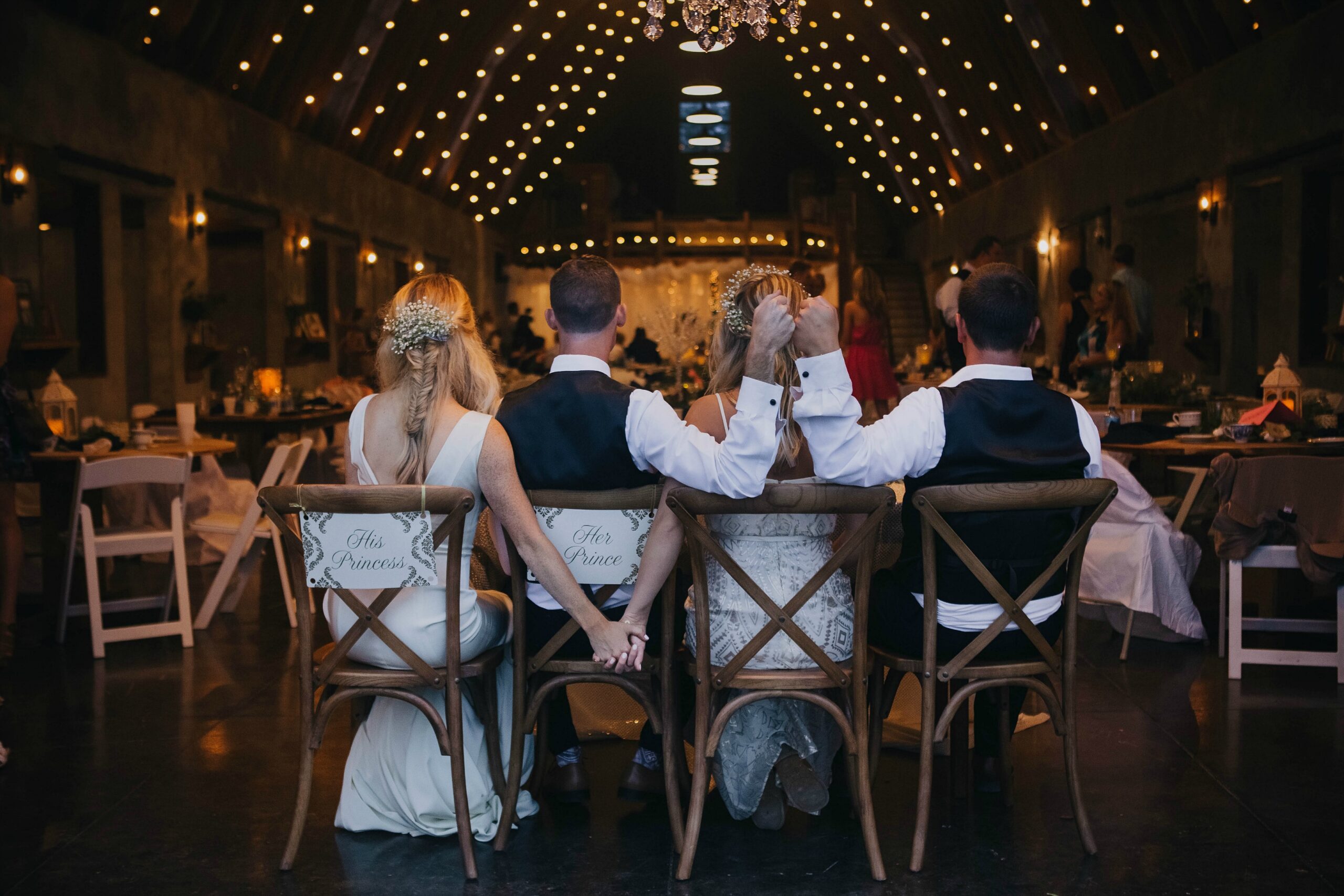 bride, groom, maid of honor, best man sit with their backs facing us and the reception in the background. Bride and bridesmaid hold hands as the men fist pump.