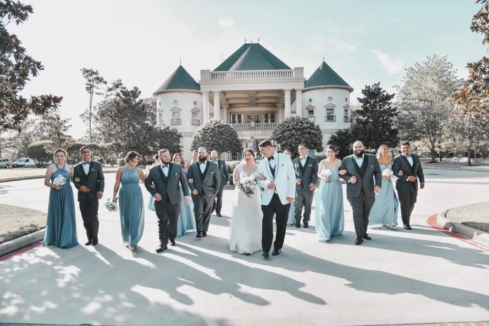 wedding party walking with a majectic building in the background. Groomsmen wearing dark grey tuxedos with groom in a white tuxedo coat and black pants. Bridesmaids in various shades of green dresses.