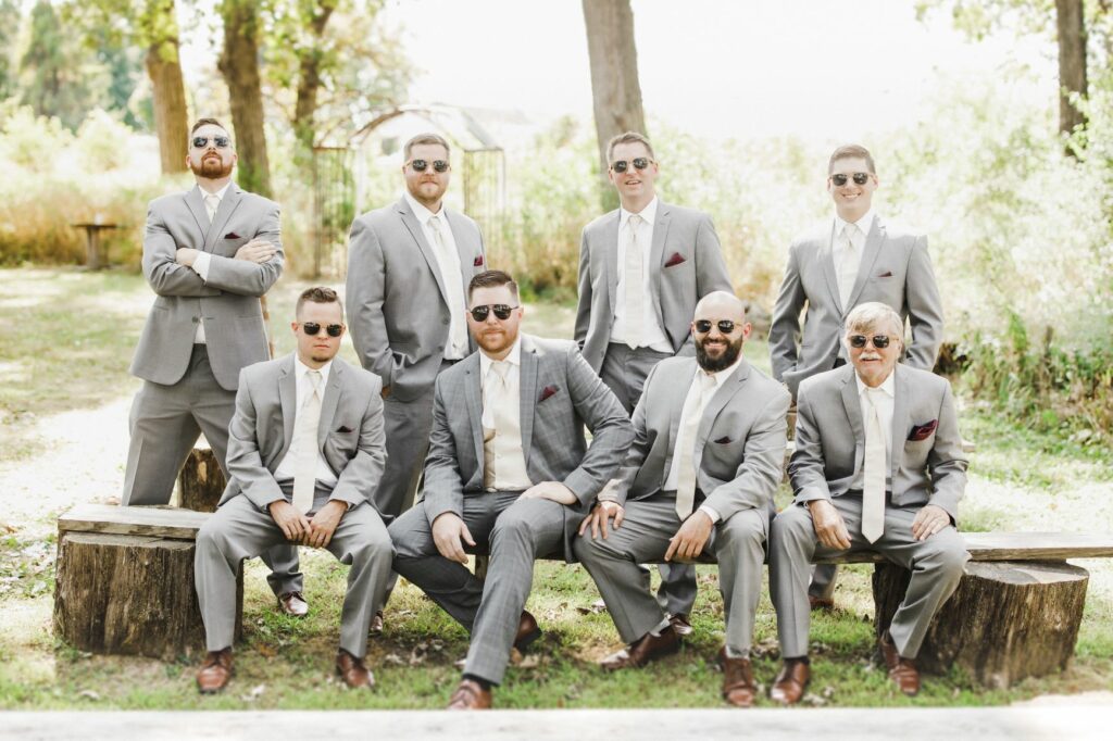 groom and groomsmen pose with sunglasses surrounded by trees. Groom in dark grey suit with groomsmen with a lighter shade of grey.