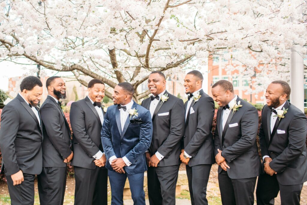 groom in navy velvet tuxedo with groomsmen gathered around him smiling and laughing and wearing black tuxedos with black accessories