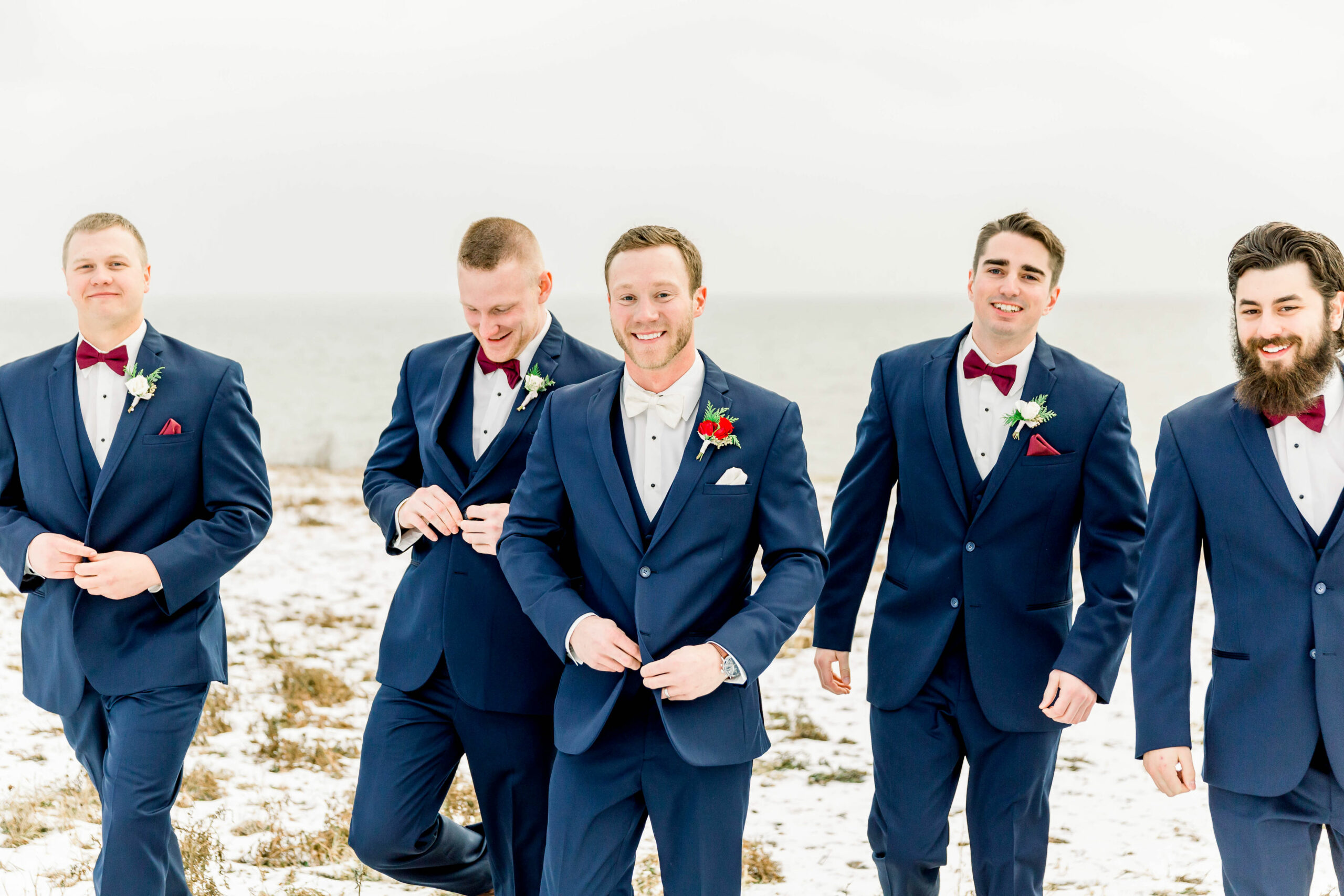 groom and groomsmen walking towards the camera in a field covered in snow. Men wearing navy blue suits with matching vests and red cranberry colored bow ties.