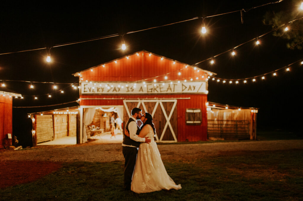 bride and groom kissing while standing in front of a red bard with string lights hanging overhead