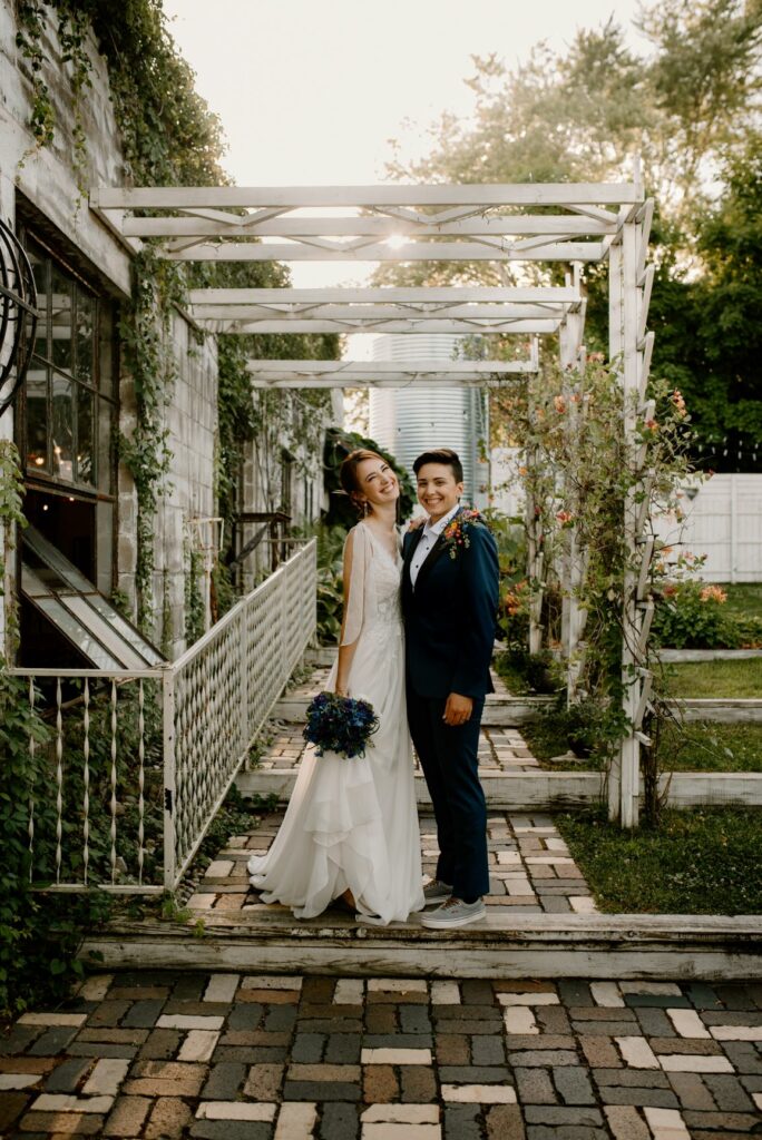 two brides standing under a trellis wrapped in flowers. One wearing a bridal dress and the other wearing a blue suit.