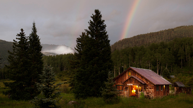 cabin nestled in the woods and hills with the steam from the hot springs rising in the background. A rainbow lights up the sky.