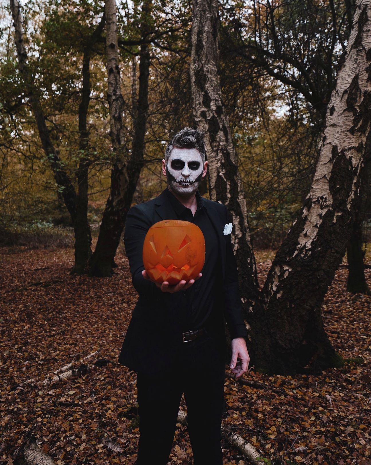 man in woods wearing all black suit with face painted like a skeleton and holding a carved pumpkin