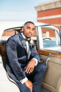 black guy in navy velvet jacket and navy pants sitting in a white classic car