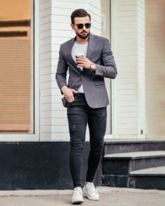 man wearing sunglass in black jeans, white t-shirt, grey suit jacket holding a cup of coffee