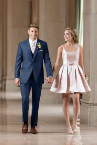 guy in blue tuxedo holding hand with a girl in a short, light pink dress