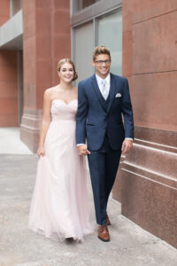 girl in long, pale pink dress holding hands with guy in blue tuxedos, walking on sidewalk