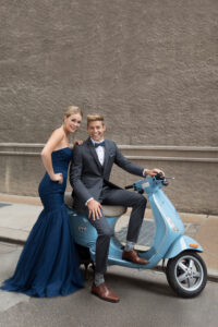 girl standing in navy blue prom dress next to a guy in a steel grey suit on a sky blue moped