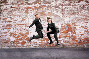 two friends jumping in black tuxedos, brick wall behind them