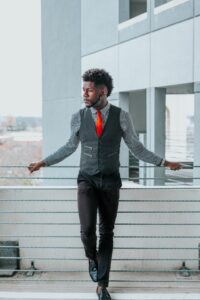 man in plaid patterned shirt, grey vest, orange tie and black jeans 