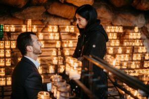 guy on on knee proposing to woman, lots of lit candles in the background