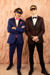 prom guy in blue tux and sunglasses, another prom guy in burgundy tux with sunglasses, they are standing in front of a feathered wall