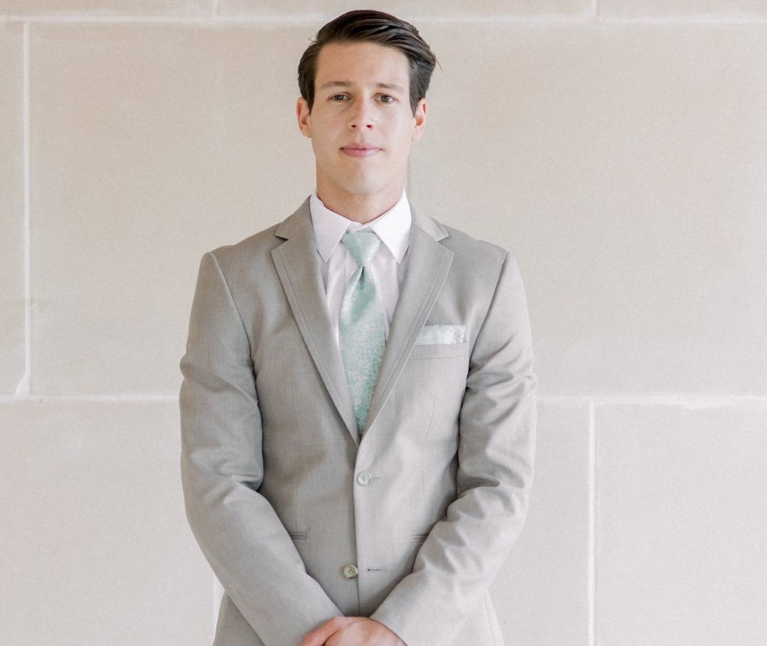 guy facing camera, wearing light tan suit and sage floral tie and pocket square. Hands are clasped together in front of him