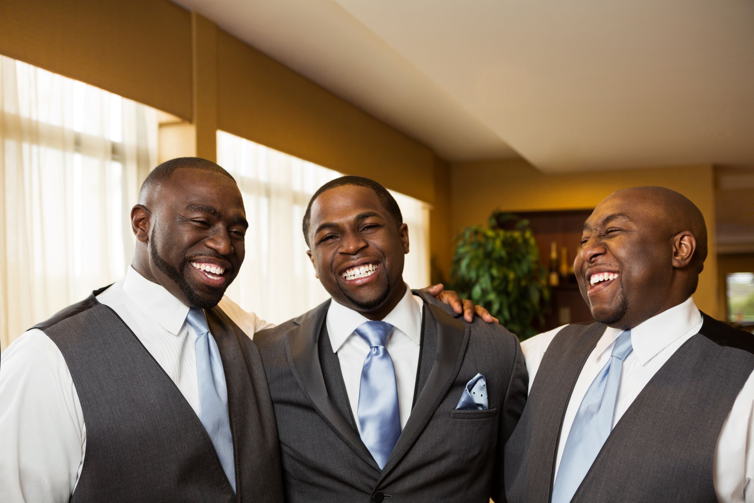 three black men standing together at a wedding. Groom is in the middle with grey tux on, each guy on the side of him are wearing grey vests and light blue ties.