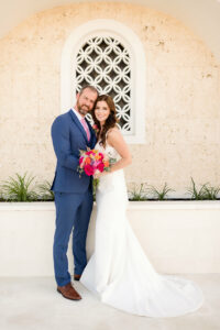 bride and groom facing camera, smiling. Groom in blue suit, bride in white wedding dress holding bouquet of bright pink and orange flowers