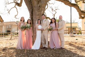 wedding party standing in line facing camera. Guys are wearing tan suit, bride in white wedding dress, bridesmaids in different shades of pink dresses