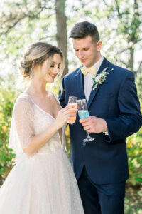 bride and groom toasting with drinks outside. Bride in white, flowy wedding dress, groom in blue suit with pale yellow bow tie