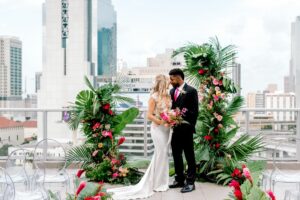 Bride and groom standing in-between large greenery floral arrangements that have bright pink flowers. Bride in white wedding dress, groom is in black suit