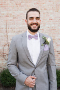guy in light grey suit with lavender bow tie, facing camera with his hand in front, holding jacket