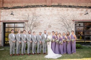 wedding party standing in line facing camera. Guys are in light grey suit , bride in white wedding dress, bridesmaids in lavender dresses 