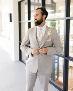 guy in light tan suit standing in front of windows, buttoning his coat at a spring wedding