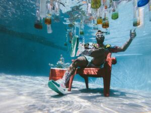 guy in a chair at the bottom of a pool with lots of liquor bottles floating above him