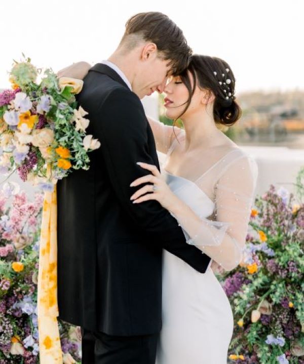 bride and groom standing together with their foreheads touching. Groom in black suit, bride in white wedding dress, holding a bouquet with lavender and yellow flowers for a spring wedding