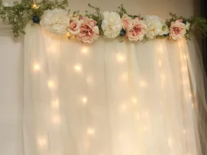 wedding backdrop with large white and pink flowers at the top, tulle underneath with fairy lights behind