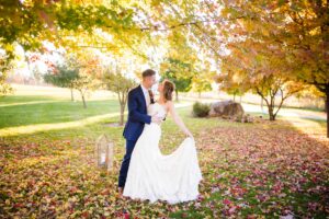 groom in navy suit with bride in white wedding dress, holding each other while outside in fall foliage 