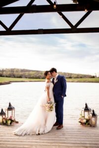 bride in wedding dress and groom in indigo blue tux standing on a dock in front of a pond 