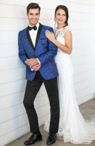 Groom in paisley cobalt blue tuxedo jacket and black pants with bride in wedding dress standing behind him with hand on shoulder