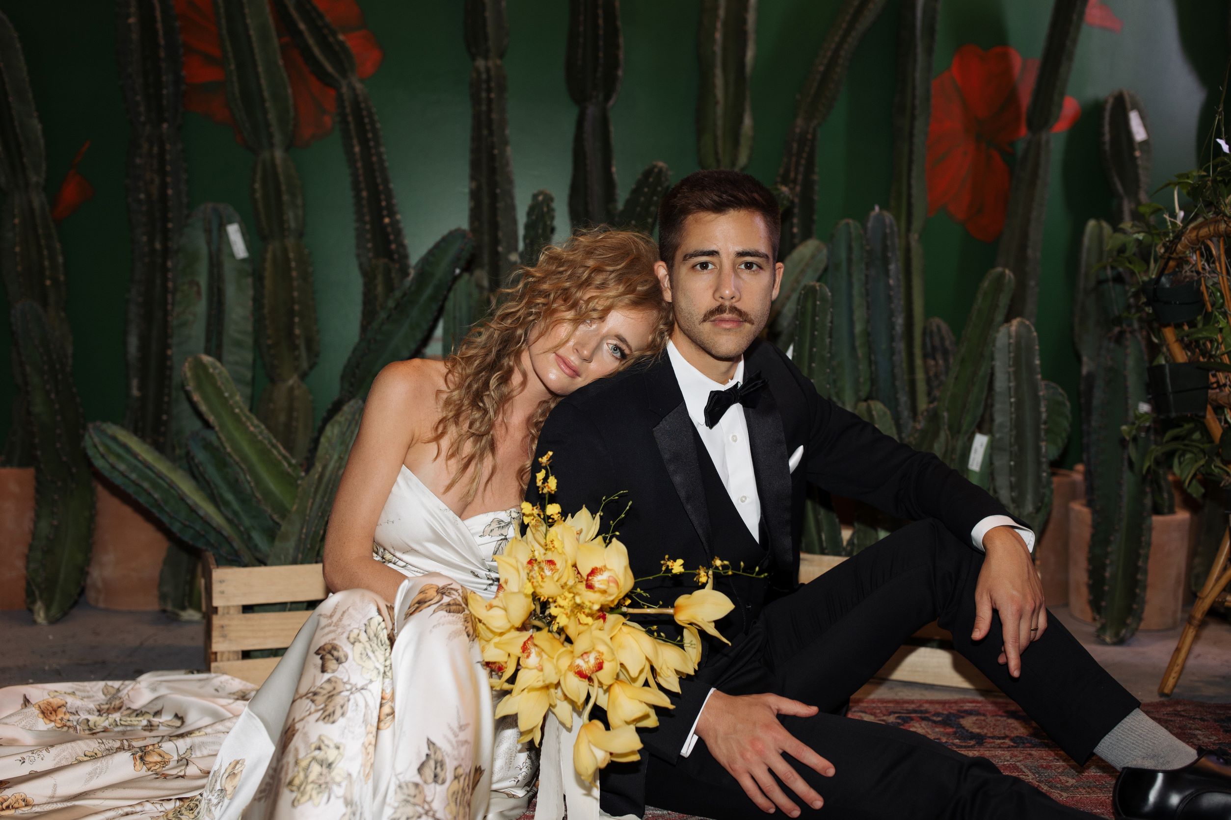 bride and groom sitting on floor, groom in black tux, bride holding bouquet of yellow flowers, behind them are cacti