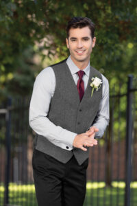 groom wearing grey tweed vest with wine tie, black pants, white shirt. White rose boutonniere pinned on left chest