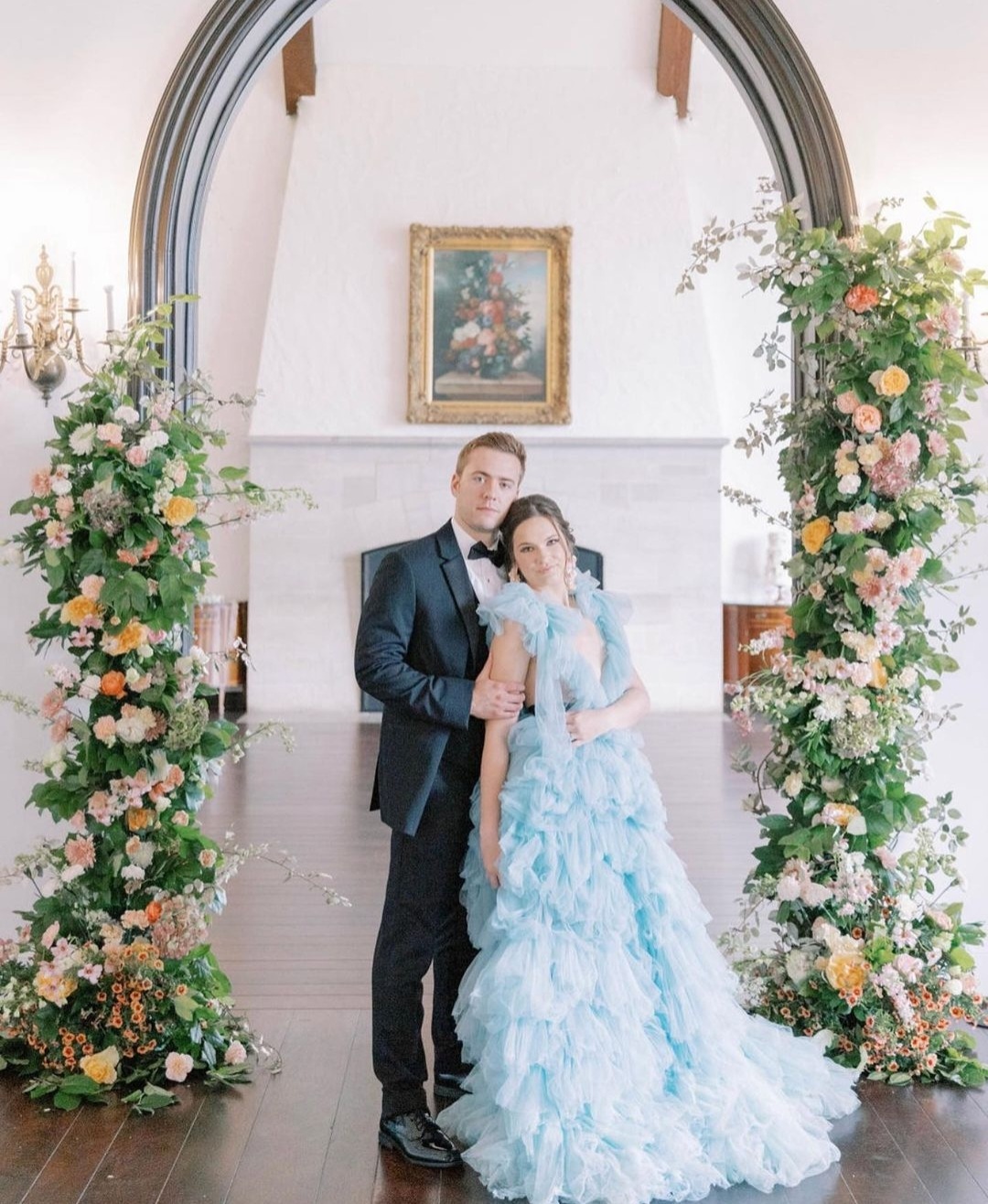 groom in black tux, standing next to bride in light blue dress. to the left and right of them are two vertical, floral arrangements