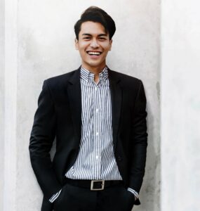 Asian guy smiling, facing camera, wearing black suit with no tie, vertical striped dress shirt