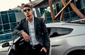 young man with sunglasses on, standing in front of grey car with door open, arm resting on window. Wearing black suit with no tie