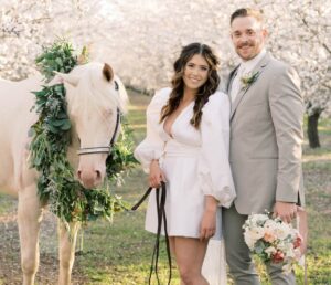 bride in short white wedding dress with puffy sleeves, standing next to groom in tan suit. bride is next to white horse that has a greenery wreath around it's neck.