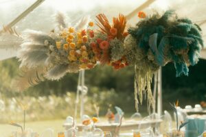 floral cloud of Pampas grass, yellow, orange roses, teal feathers, baby's breath above wedding reception table. 
