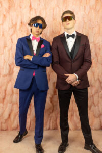 high school guys standing in front of light pink feather wall. One boy is in cobalt blue tuxedo with pink bow tie, vest and pocket square. Other boy is in burgundy jacket with black pants, black bow tie
