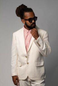 black guy with dreads in a bun, wearing sunglass, white suit with no tie, pink button down shirt. His hand is at his chin