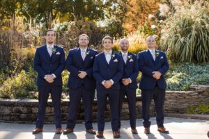 groom and groomsmen standing in line wearing navy suits, hands are held in the front of them